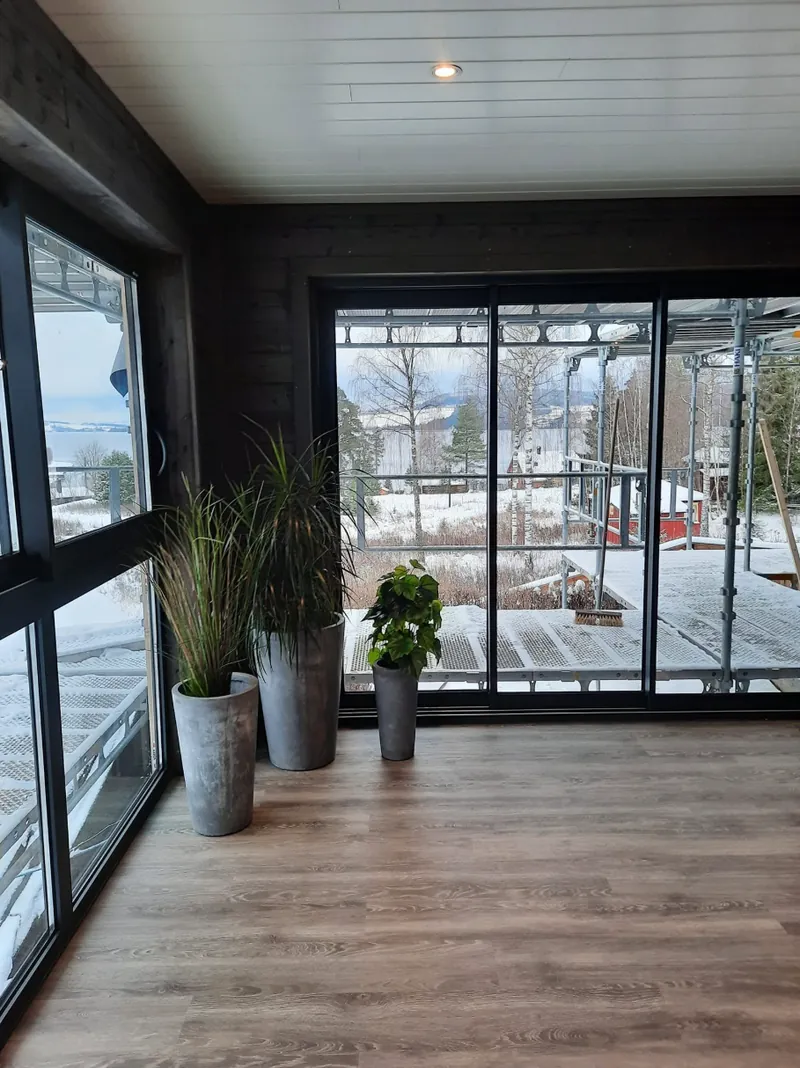 Hagestue med store glassfelt, mørk panel og planter, med utsikt over snødekt landskap