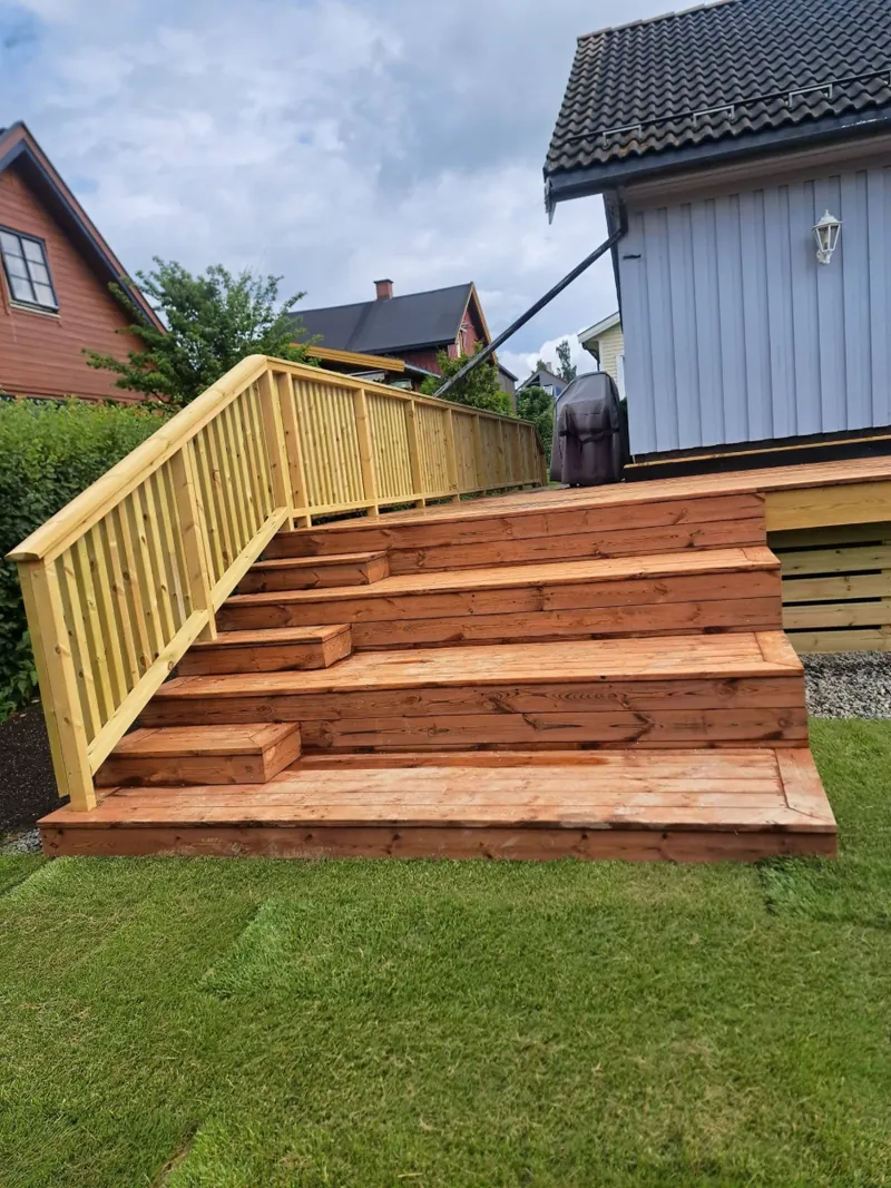 Bred trapp i terrassebord opp til ny terrasse med rekkverk, foran et lyseblått hus